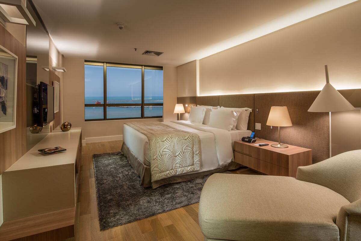 A hotel room with a bed, bedside table, lamp, chair, and a large window with a view of the sea.