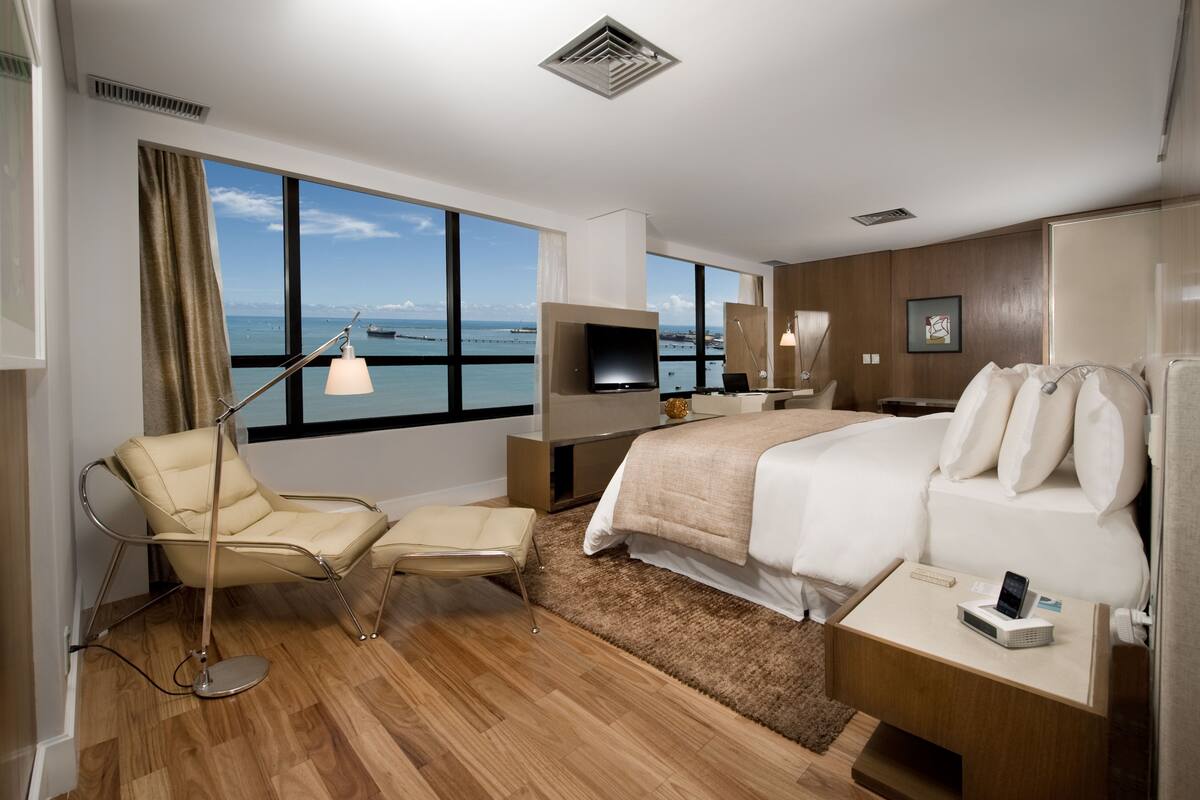 A modern hotel room with a large window, a bed, a desk, a chair, and a TV.