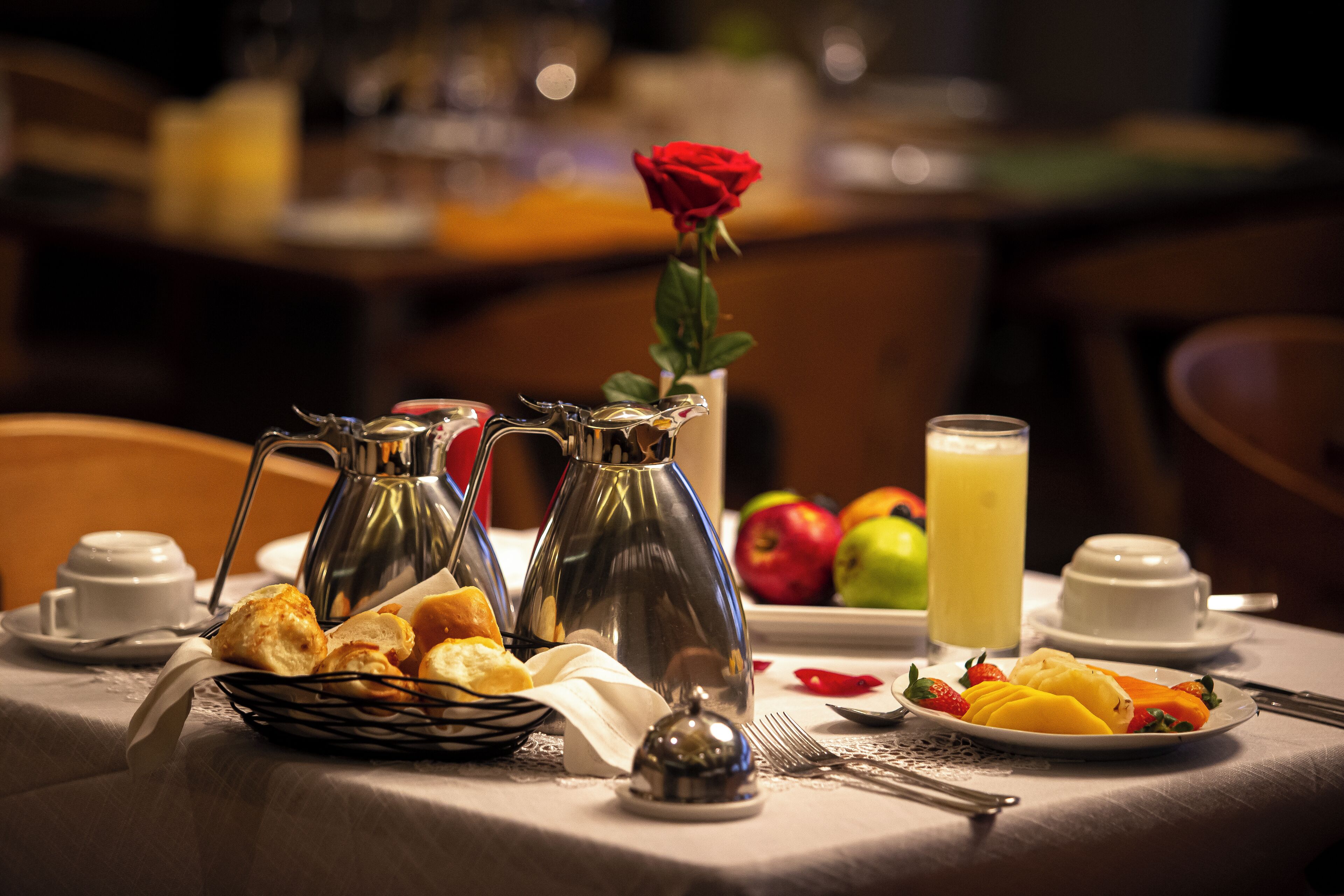 A breakfast table with coffee pots, a rose, fruits, and a glass of juice.