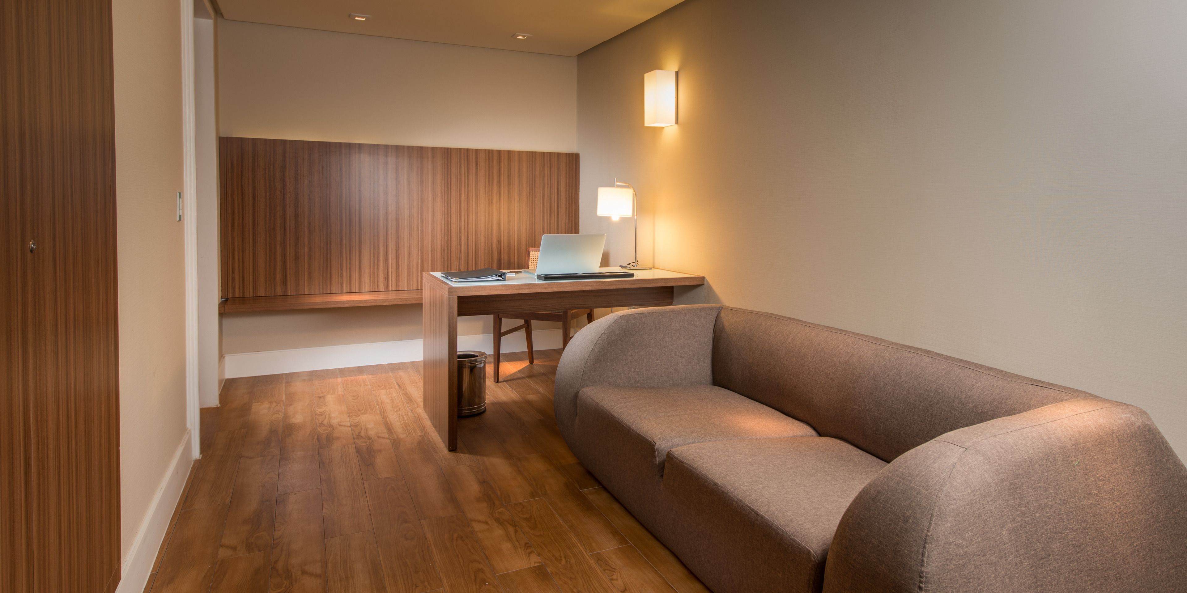 Una habitación moderna con suelo de madera, un sofá gris, un escritorio con una computadora portátil y una lámpara de pared.