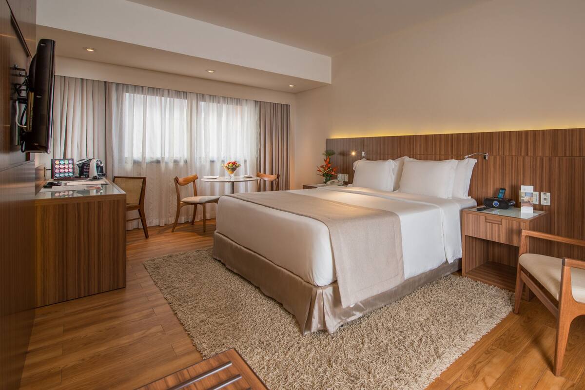 A modern hotel room with a large bed, a desk with a chair, a TV, and a small table with a vase of flowers.
