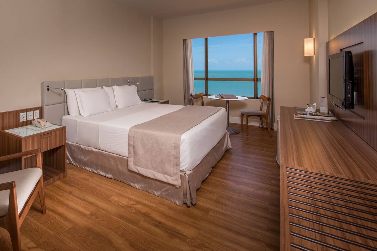 A hotel room with a bed, a desk, a chair, a TV, and a view of the ocean.