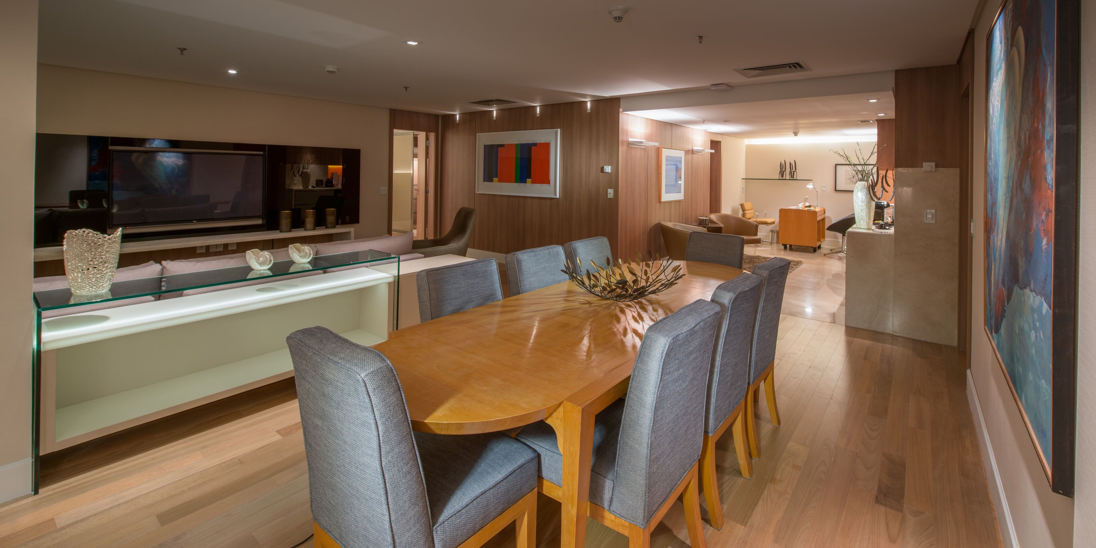 A modern dining area with a wooden table and grey upholstered chairs, a glass-top bar counter, and a flat-screen TV mounted on the wall.