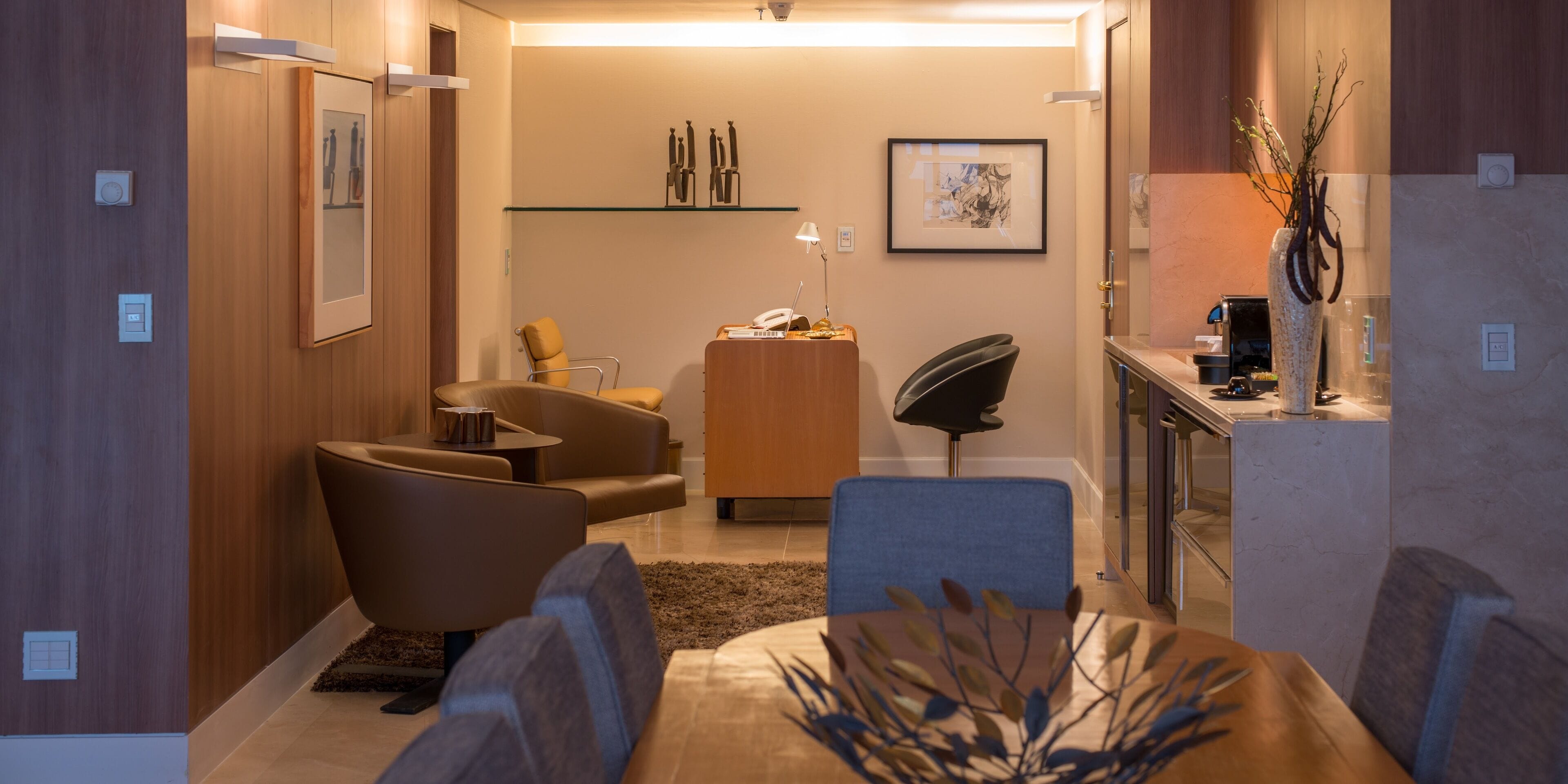 A modern living room with a sofa, armchairs, a coffee table, and a dining table. There is a small kitchen area with a coffee machine and a vase with branches.