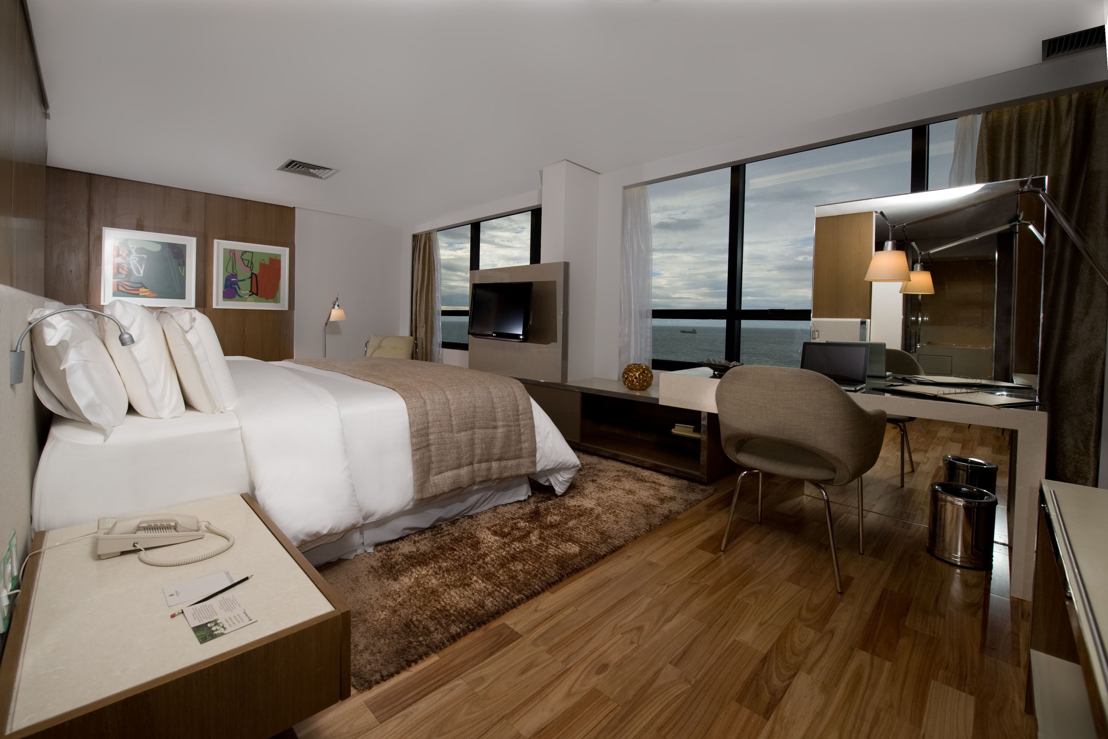 Ein modernes Hotelzimmer mit einem großen Bett, einem Schreibtisch, einem Sessel, einem Fernseher und Blick aufs Meer.