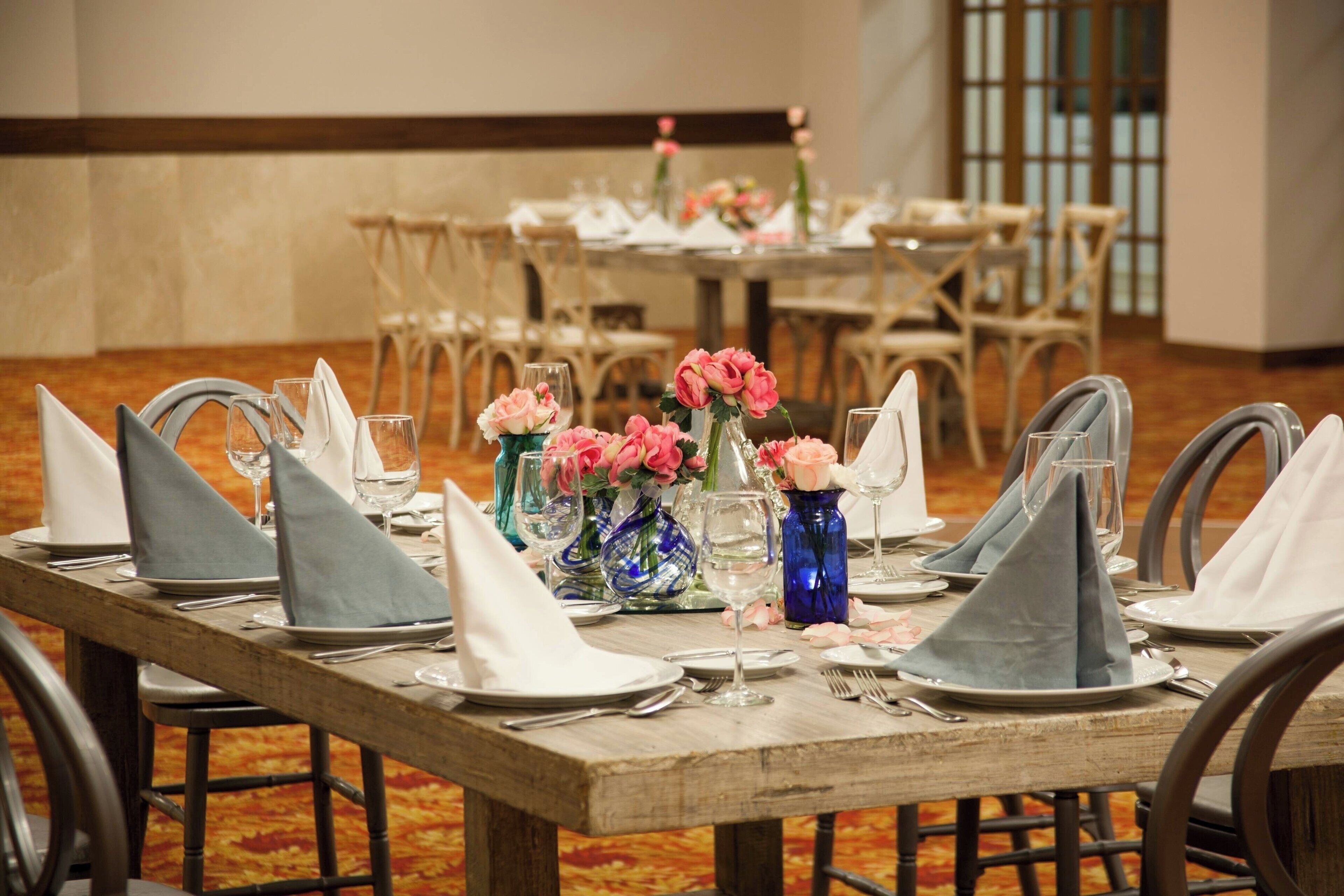 Una mesa de banquete dispuesta con servilletas azules, platos blancos y flores rosas en jarrones.