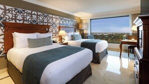 A hotel room with two beds, a desk, a chair, and a large window offering a city view.