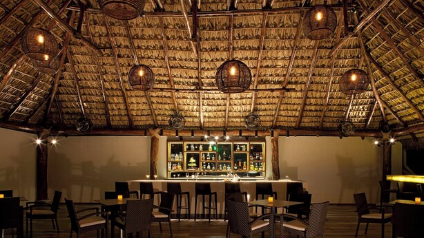 An indoor dining area with a thatched roof, wicker furniture, and a bar with a variety of bottles.