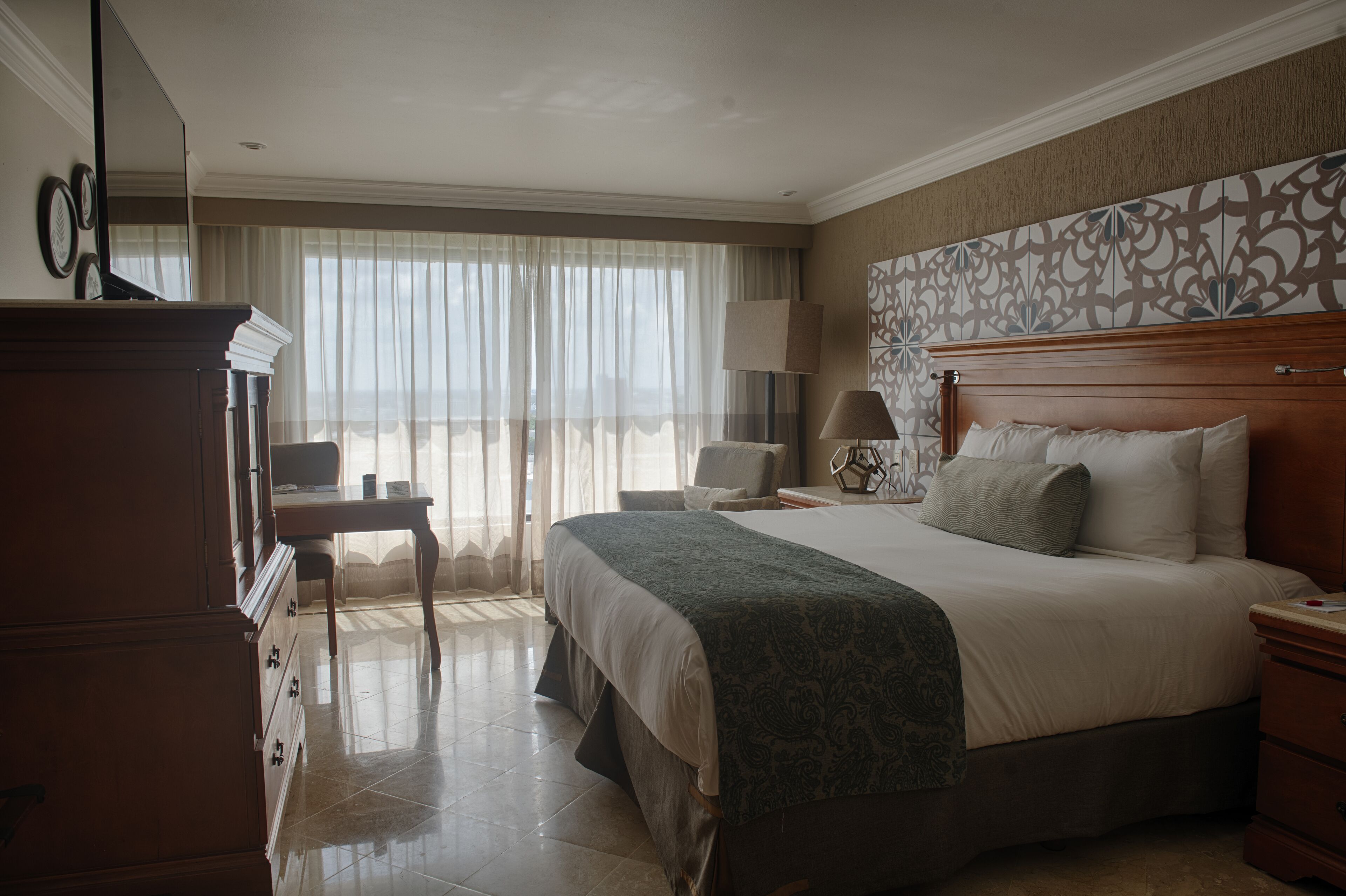 Habitación de hotel con una cama grande, un escritorio, una silla, un aparador y una ventana con cortinas traslúcidas.