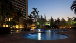 A swimming pool with lounge chairs and palm trees, surrounded by a building with multiple floors.
