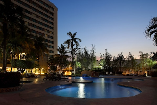 Una piscina con sillones de descanso y palmeras, rodeada por un edificio de varios pisos.