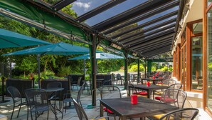 Un patio al aire libre con mesas y sillas, cubierto por un techo de vidrio y rodeado de vegetación.