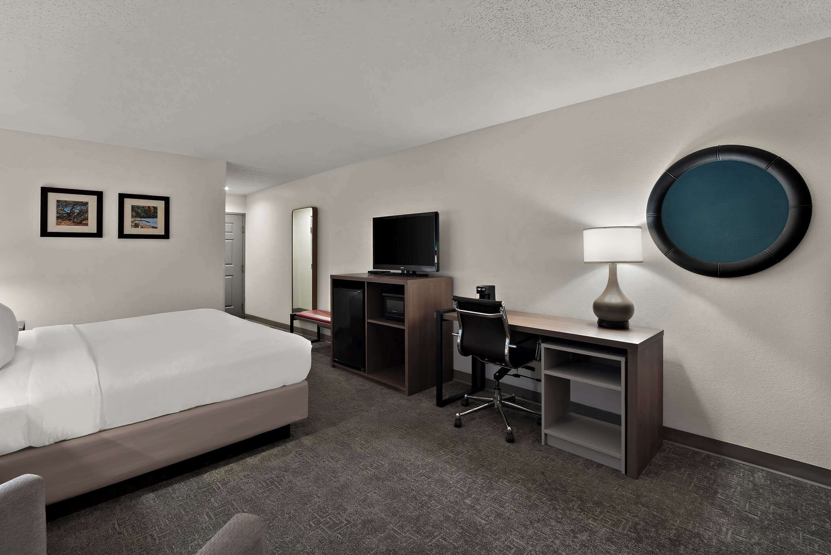 Une chambre d’hôtel avec un lit, un bureau, une chaise, une télévision et un miroir rond accroché au mur.