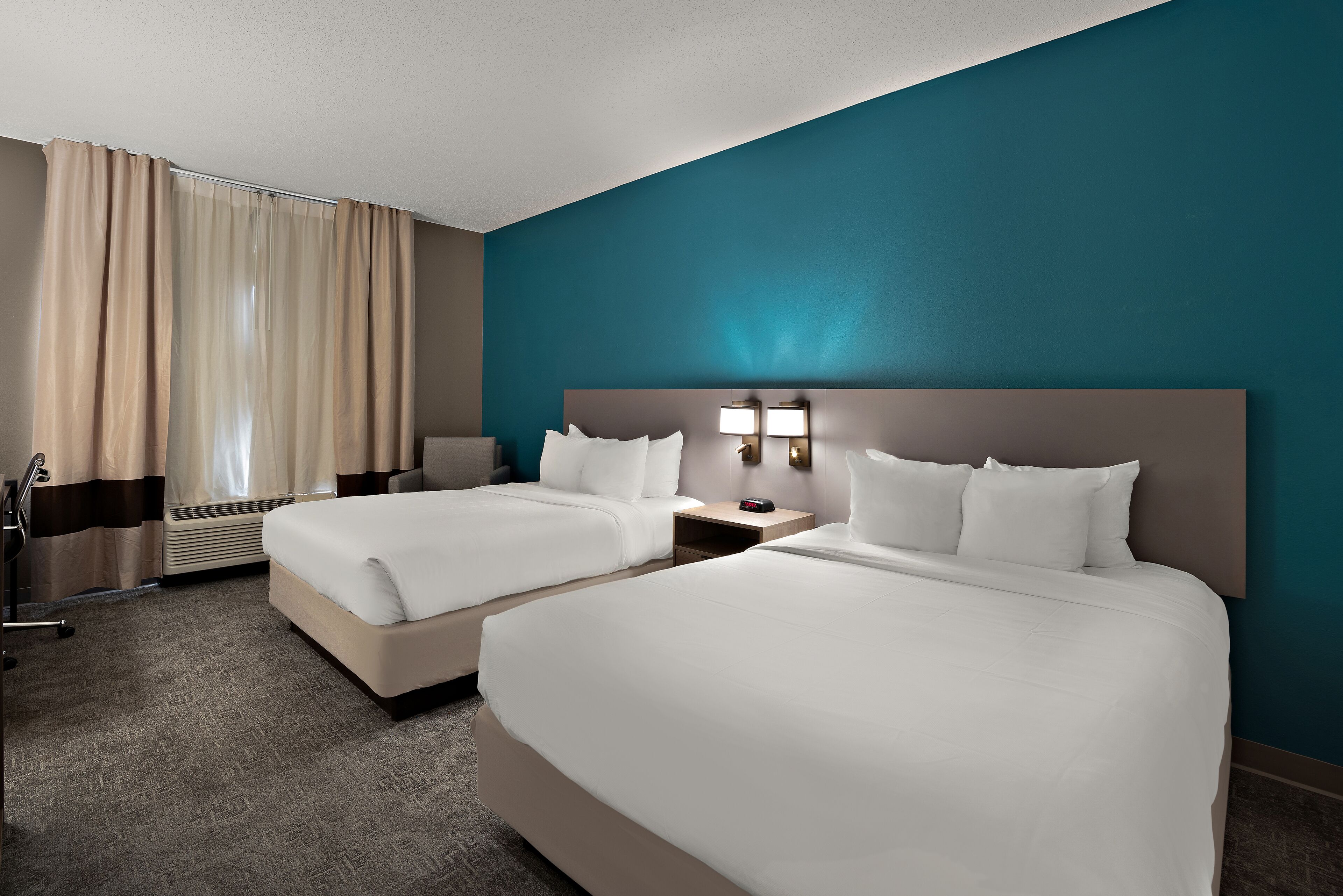Ein Hotelzimmer mit zwei Betten, einem Schreibtisch, einem Stuhl, einer Lampe und einem Fenster mit Vorhängen.