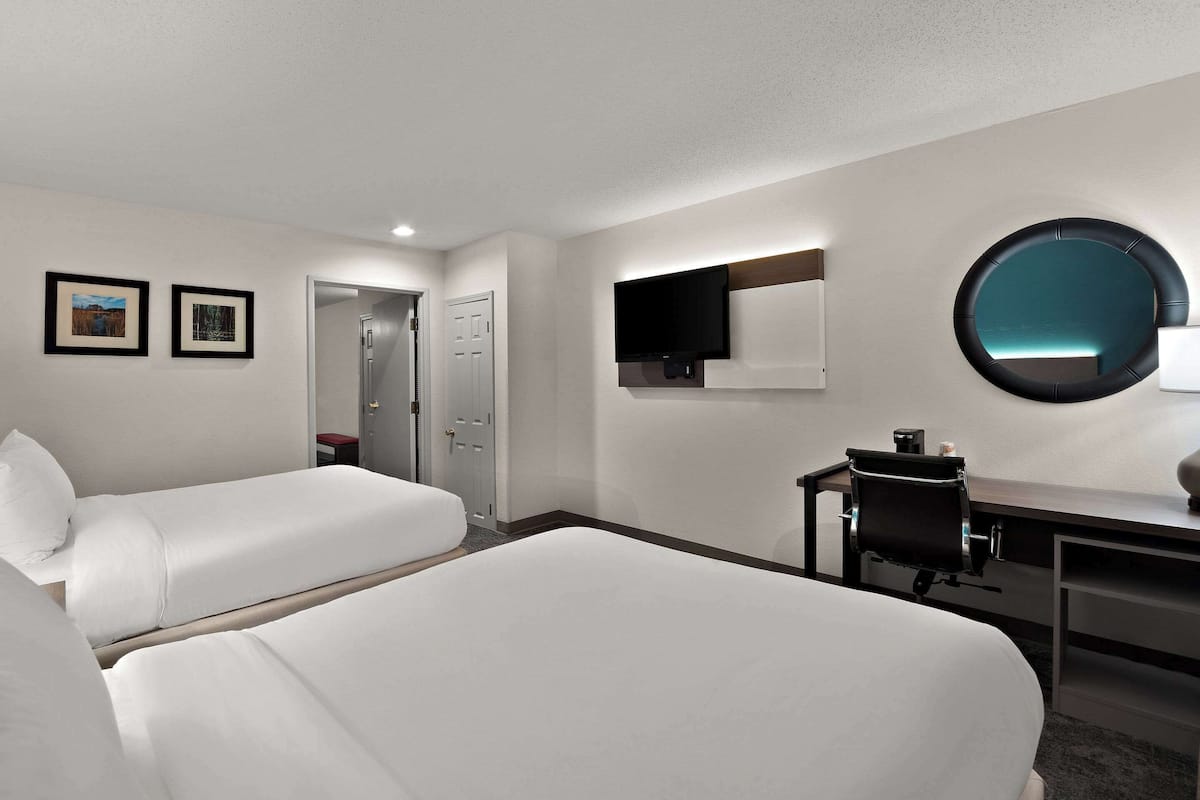 Una habitación de hotel con dos camas individuales, un escritorio con silla, un espejo redondo y un televisor de pantalla plana montado en la pared.