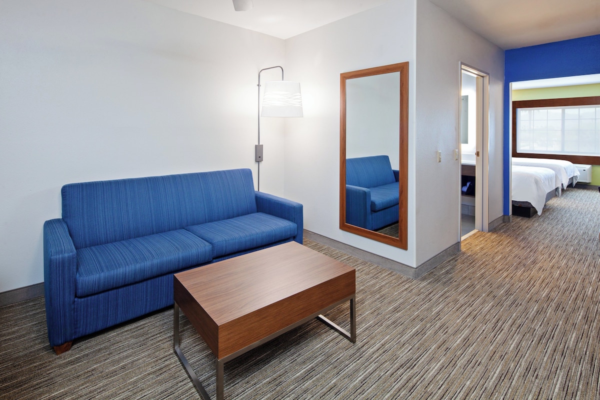 Une chambre d’hôtel avec un canapé bleu, une table basse en bois, un miroir et un lit recouvert de draps blancs.