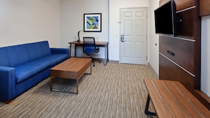 A modern room with a blue sofa, a wooden desk, a chair, a television mounted on a wooden panel, and a door. - Holiday Inn Express & Suites Rancho Mirage - Palm Spgs Area by IHG (Rancho Mirage)