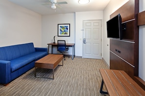 Une pièce moderne avec un canapé bleu, un bureau en bois, une chaise, un téléviseur fixé sur un panneau en bois et une porte.