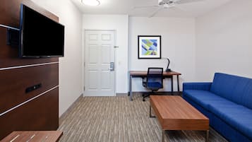Een moderne hotelkamer met een flatscreen televisie aan de muur, een houten bureau met een stoel, een blauwe bank en een plafondventilator.
