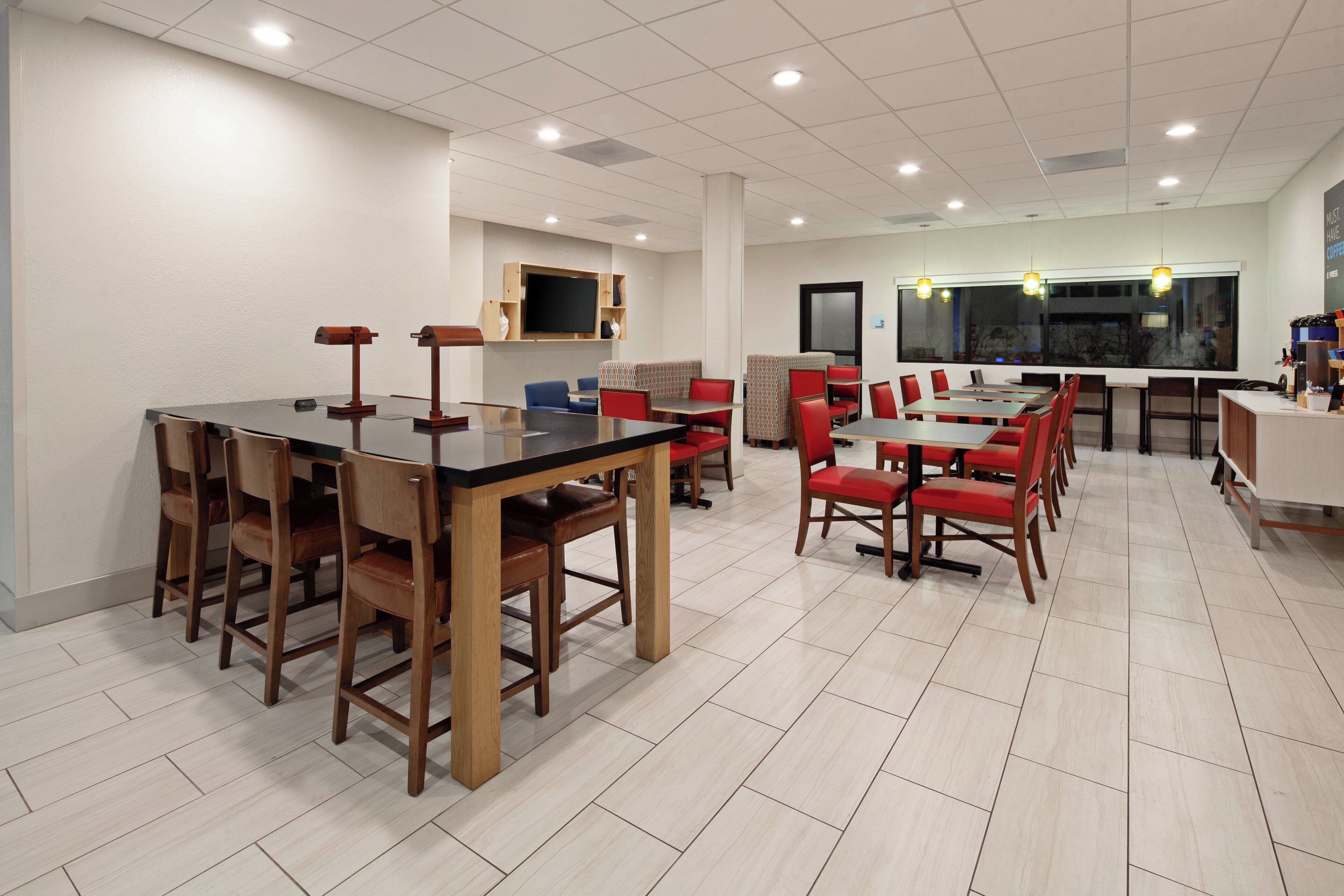 Un espace repas moderne avec des tables et des chaises en bois, un téléviseur à écran plat fixé au mur et une grande fenêtre offrant une vue sur l’extérieur.