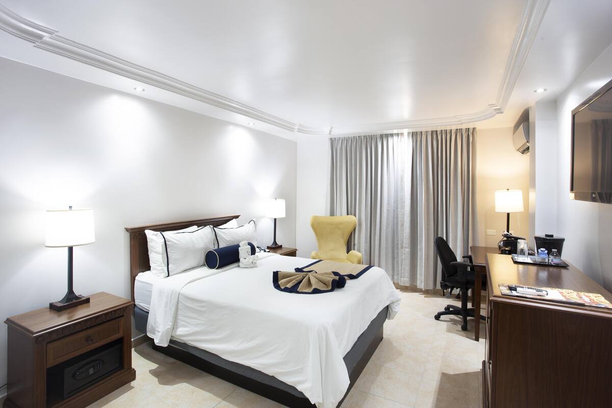 A hotel room with a neatly made bed, a desk with a chair, a lamp, and a television.