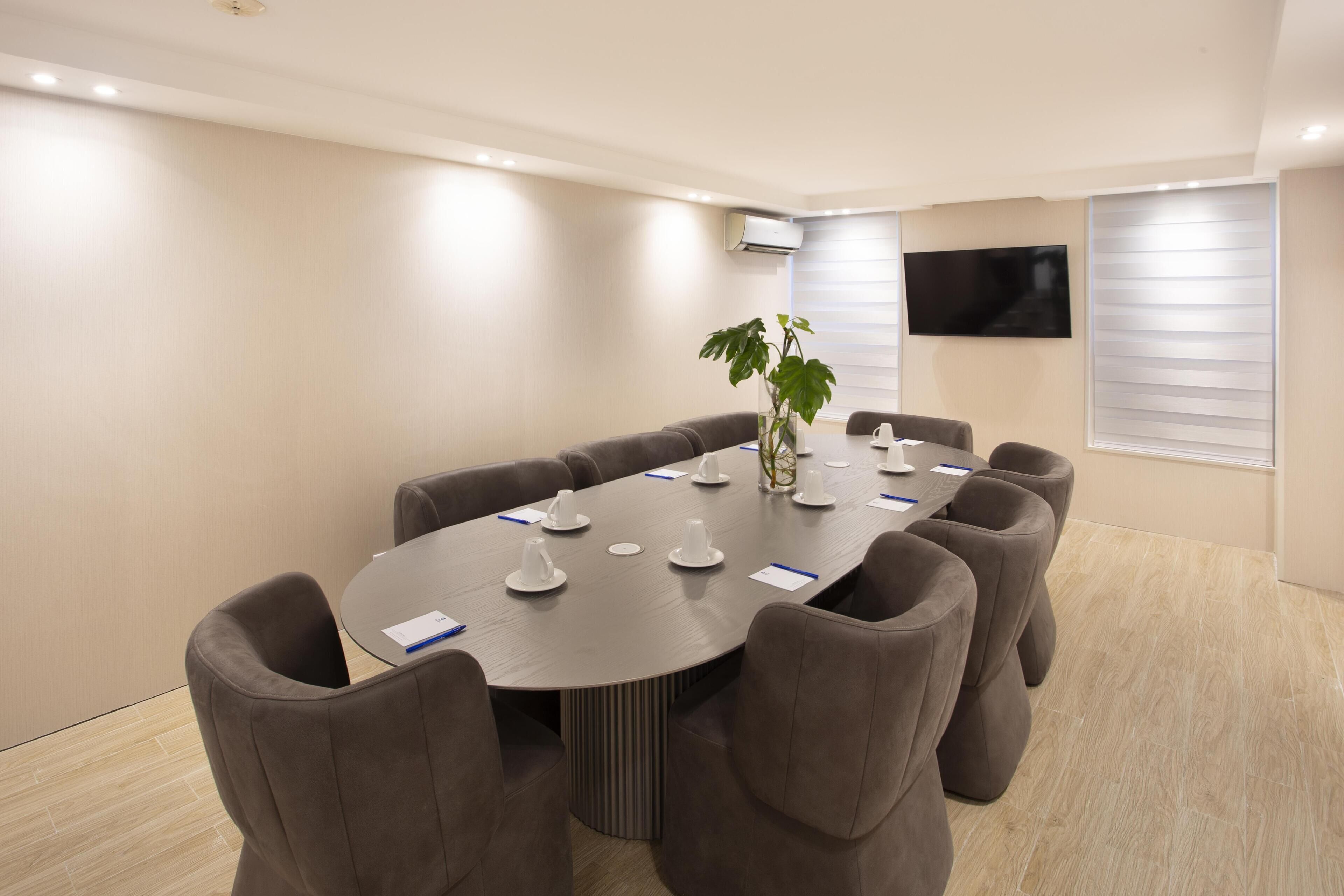 A conference room with a large oval table, eight upholstered chairs, a potted plant, and a flat-screen TV mounted on the wall.