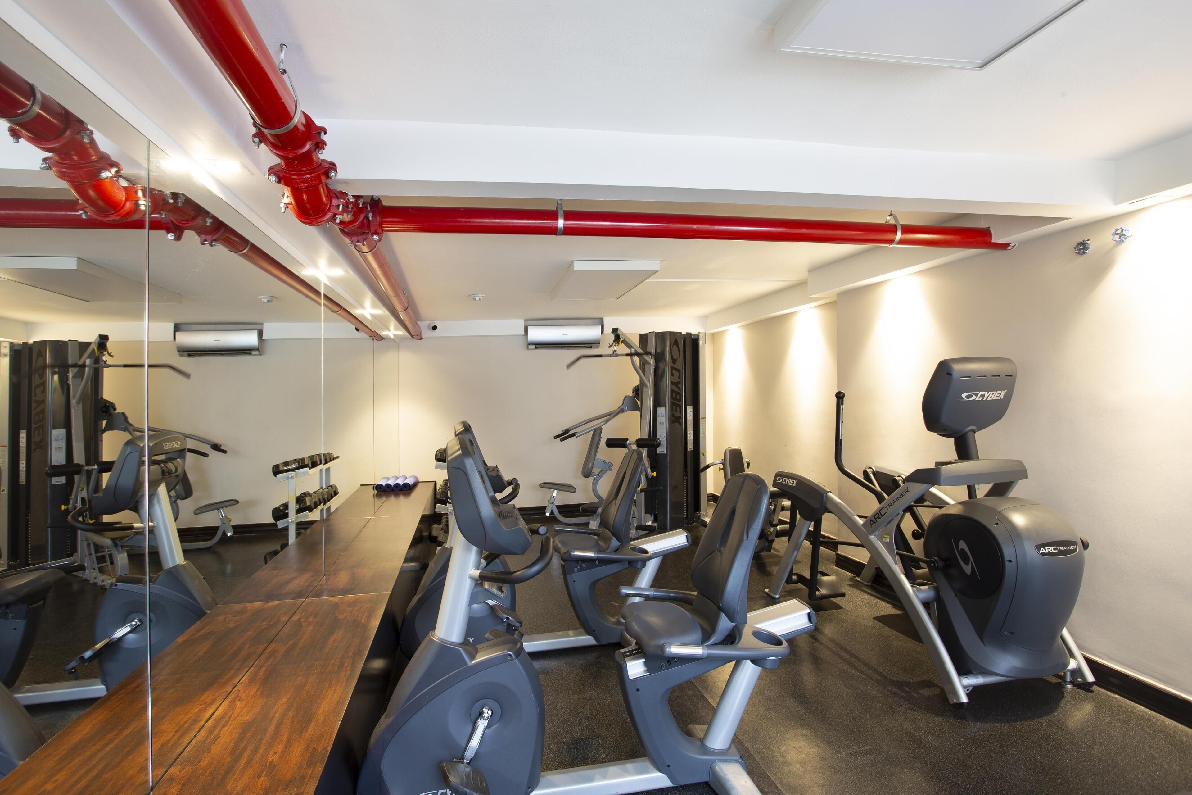 Uma academia com equipamentos de exercício, piso de madeira e um cano vermelho no teto.
