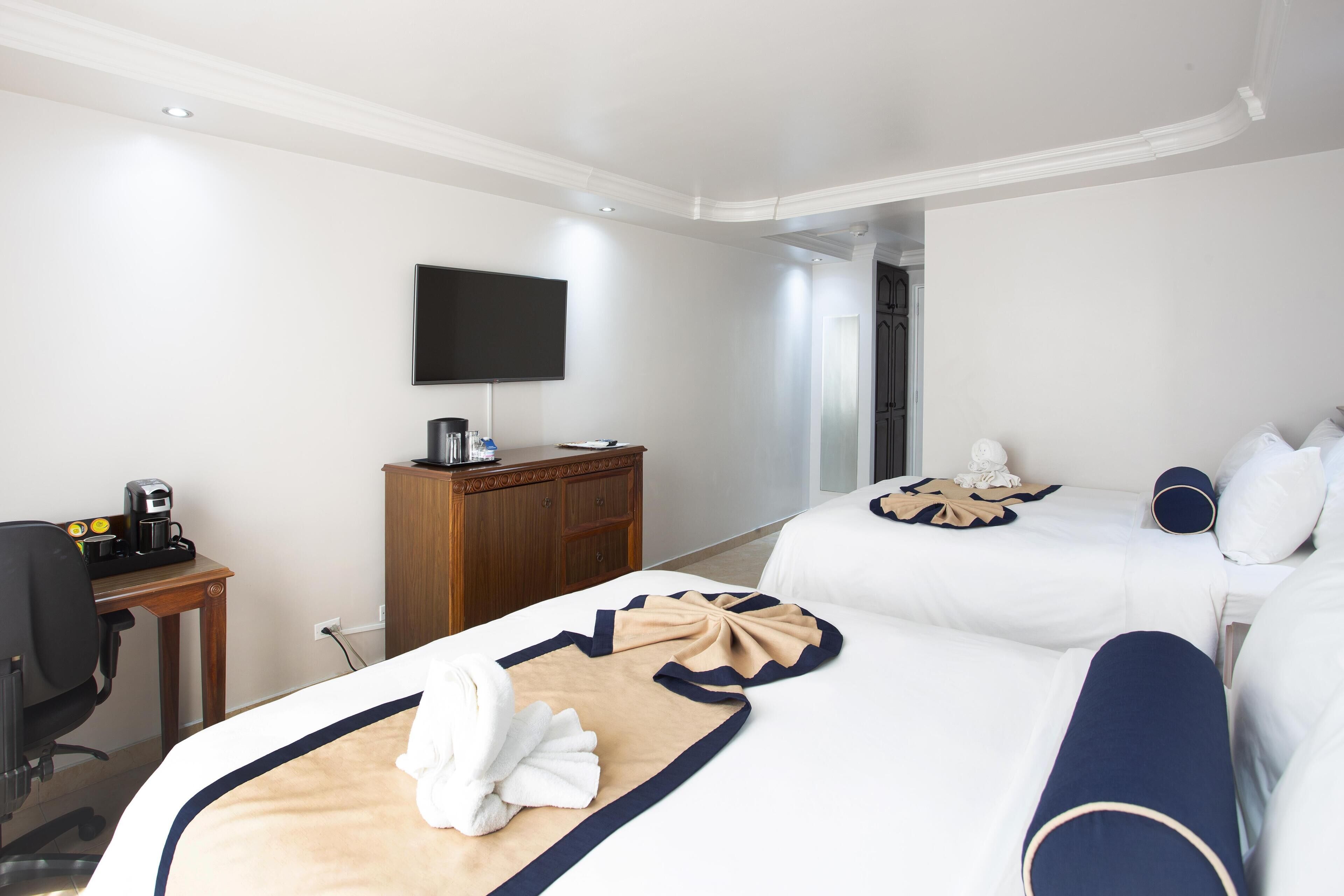 Quarto de hotel com duas camas, uma escrivaninha com cafeteira, televisão na parede e um guarda-roupa.