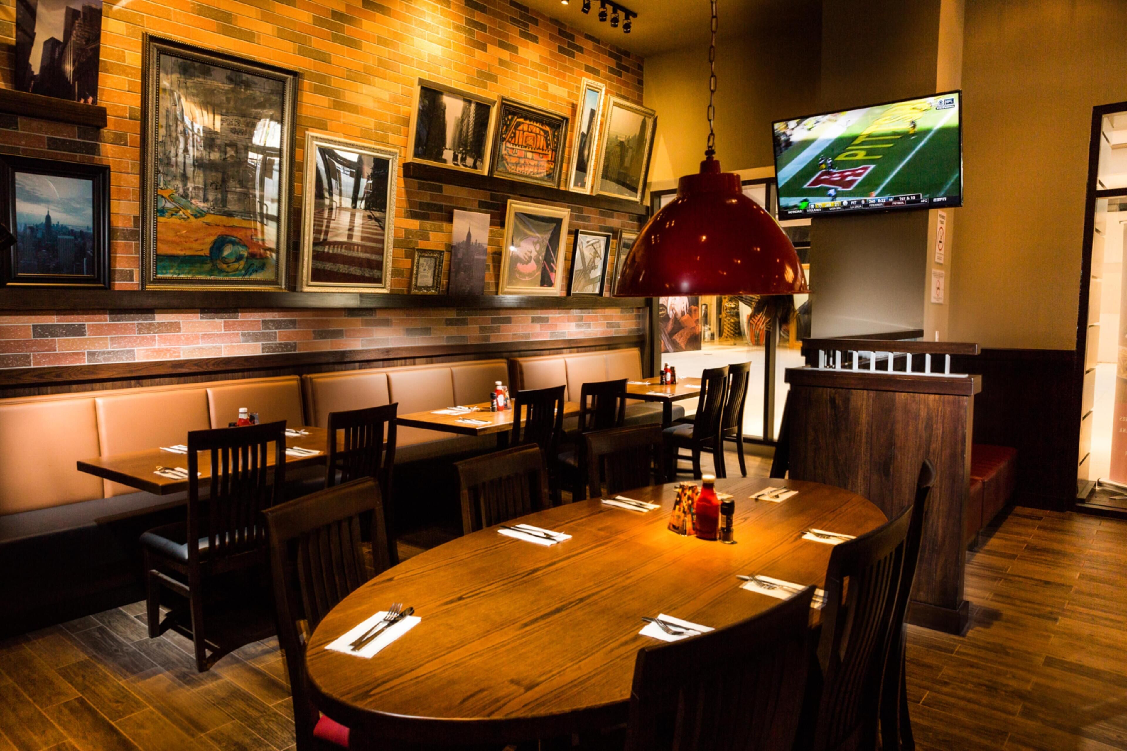 Un restaurant aux murs de briques, décoré de tableaux encadrés, avec une grande table en bois et un téléviseur diffusant un événement sportif.