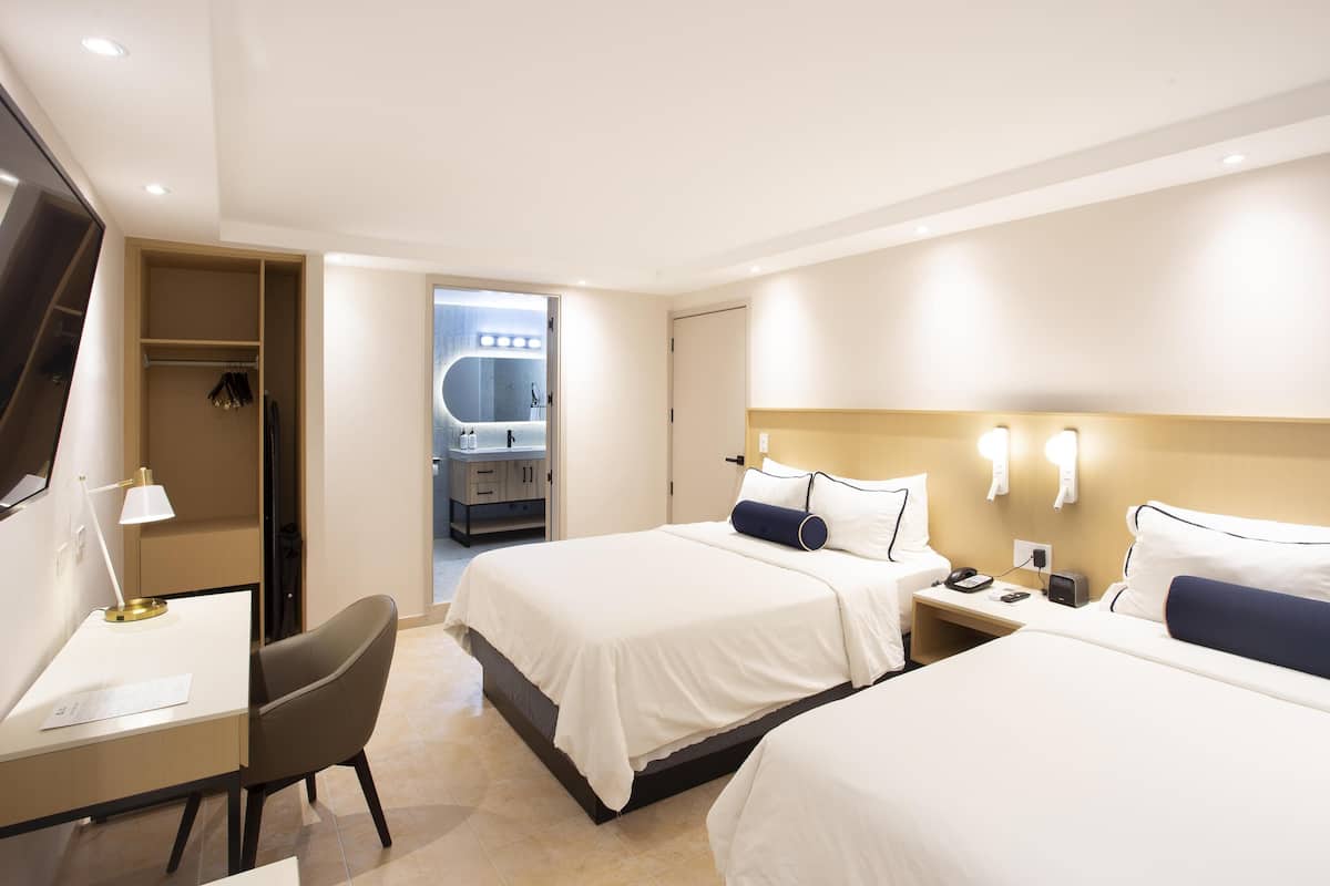 A hotel room with two beds, a desk, a chair, a TV, and a bathroom visible through an open door.