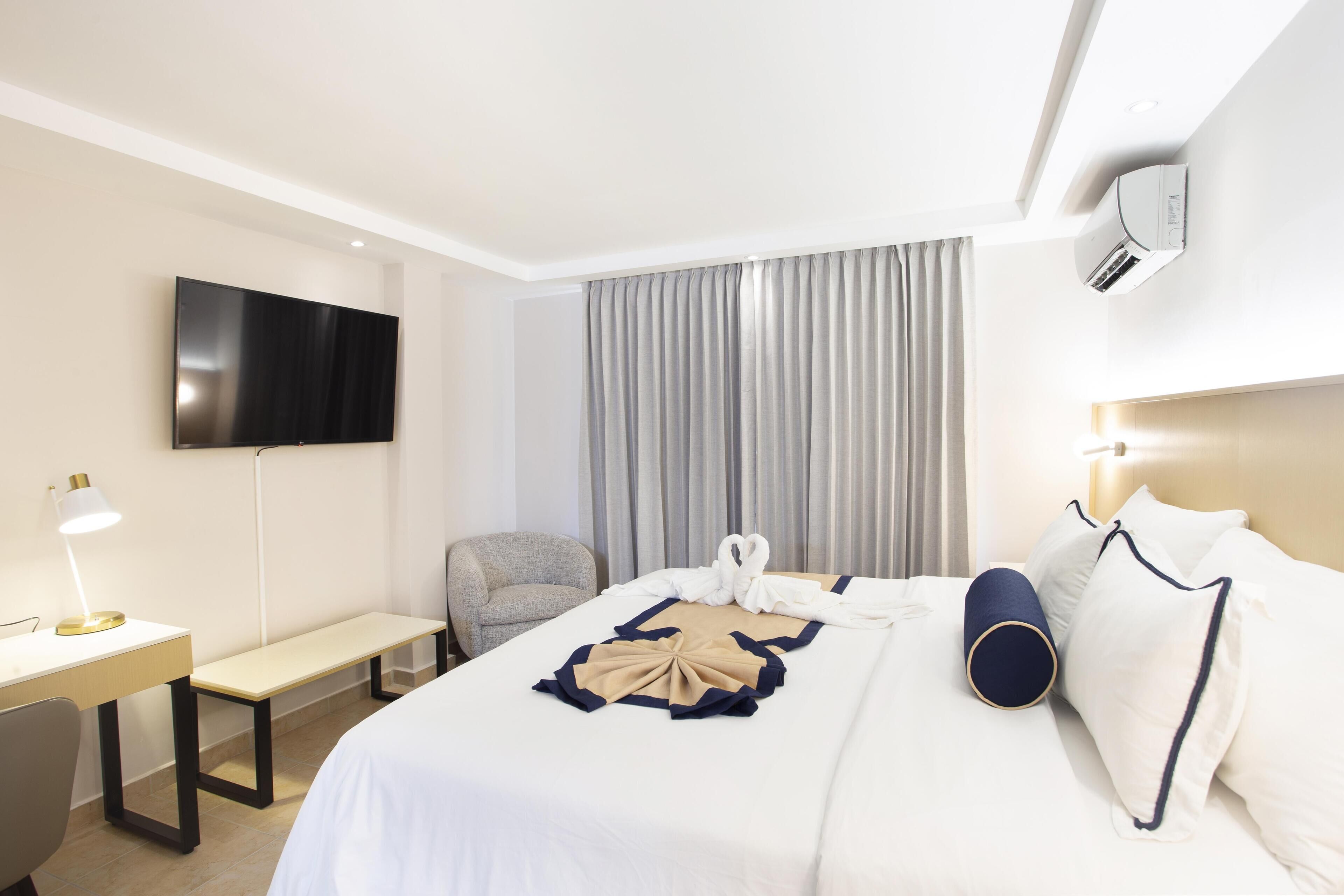 Ett hotellrum med en säng, ett skrivbord, en stol, en tv och ett fönster med gardiner.