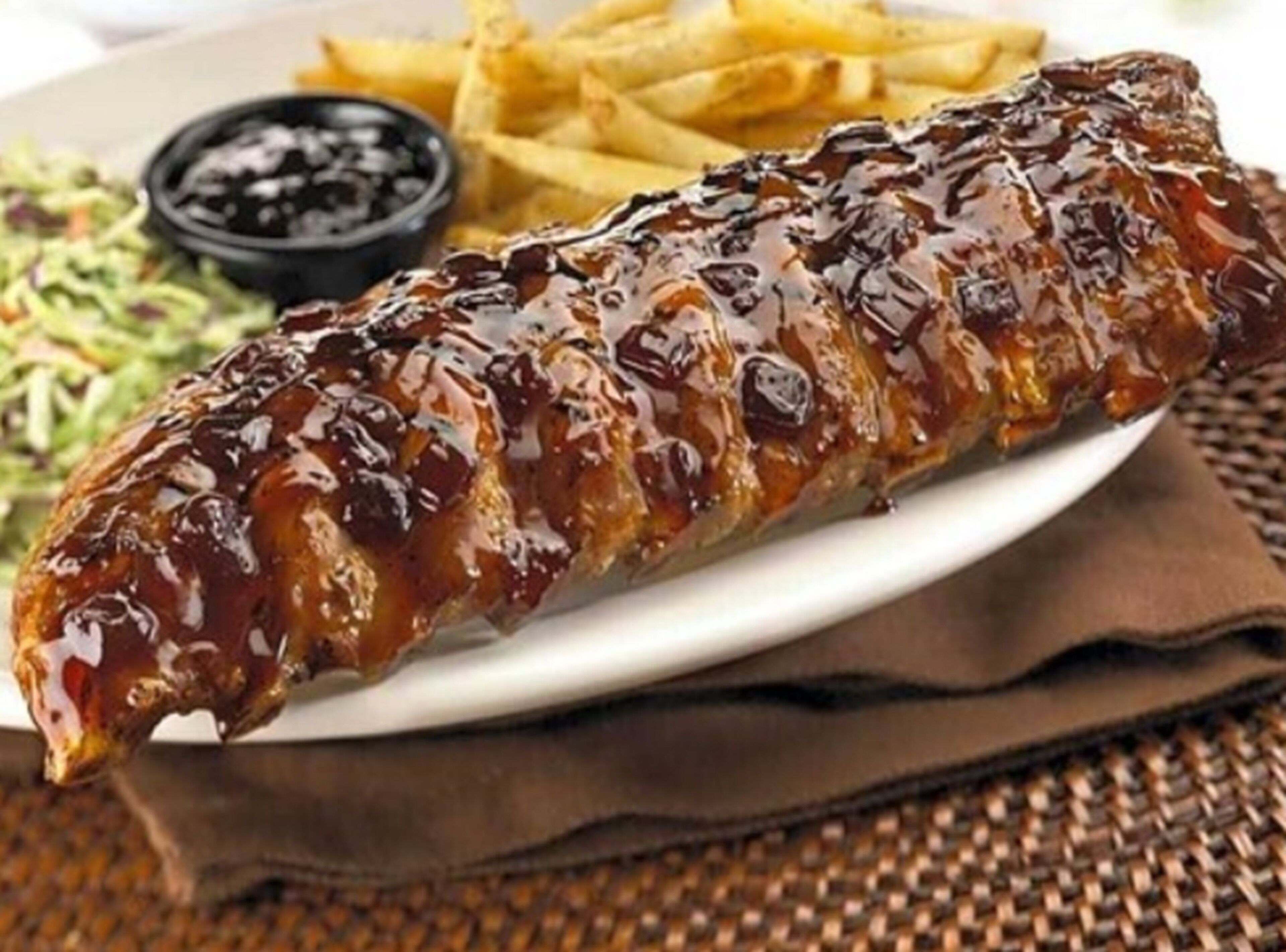 Un plato con costillas a la parrilla, papas fritas, ensalada de col y un recipiente pequeño con salsa.