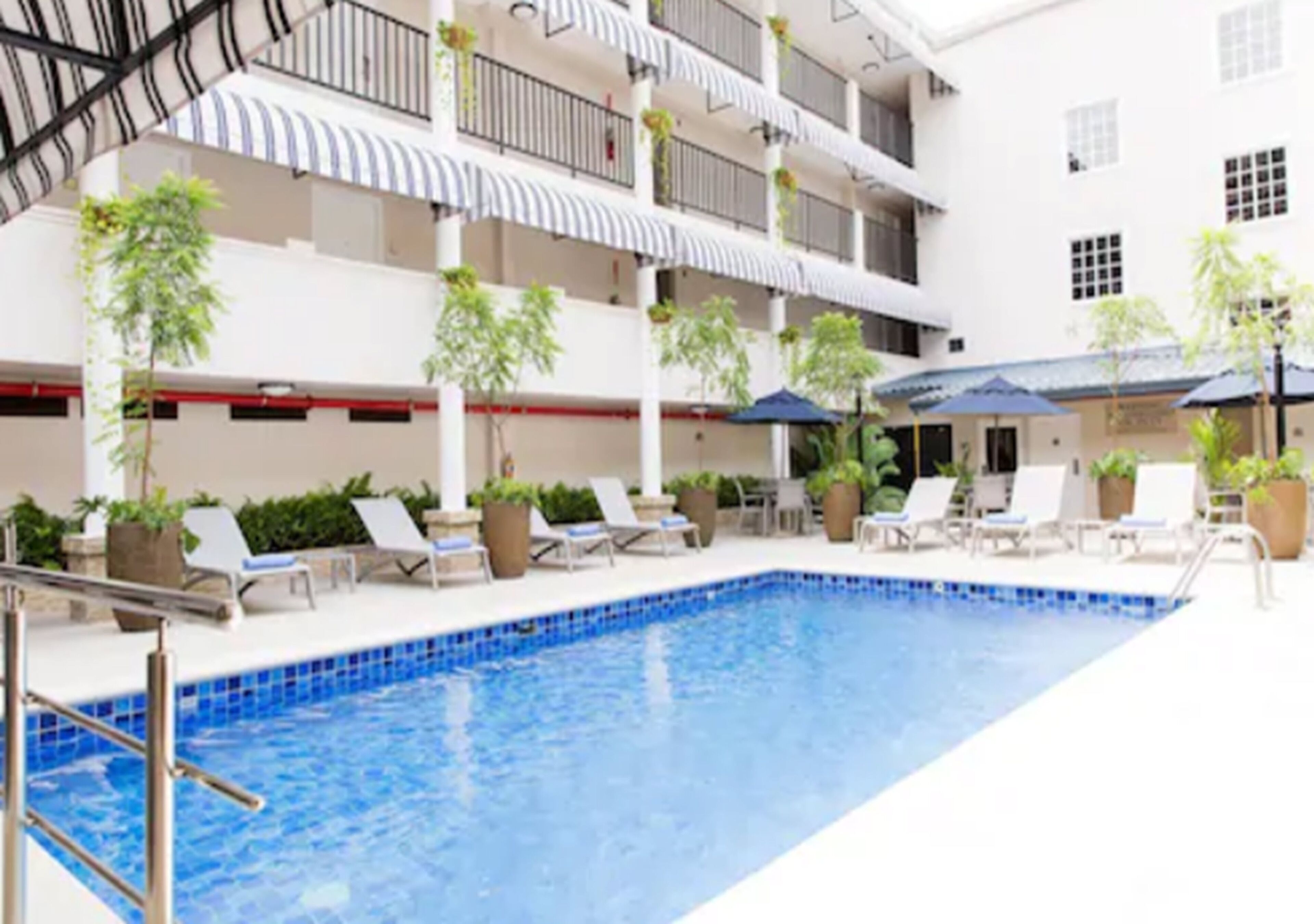 An indoor swimming pool with lounge chairs and umbrellas, surrounded by potted plants and a multi-level building with balconies.