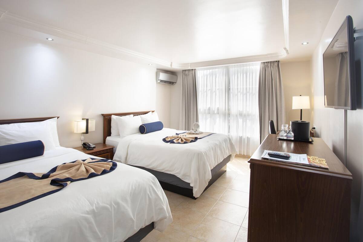 A hotel room with two beds, a desk, a TV, and a window with curtains.