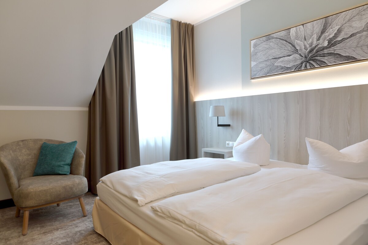 Una habitación de hotel con una cama grande, una silla, una lámpara y una obra de arte en la pared.