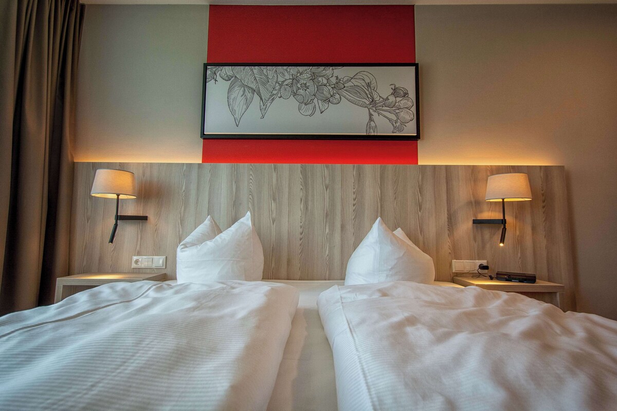 Habitación de hotel con dos camas, cabecero de madera, pared de acento roja y lámparas fijadas a la pared.