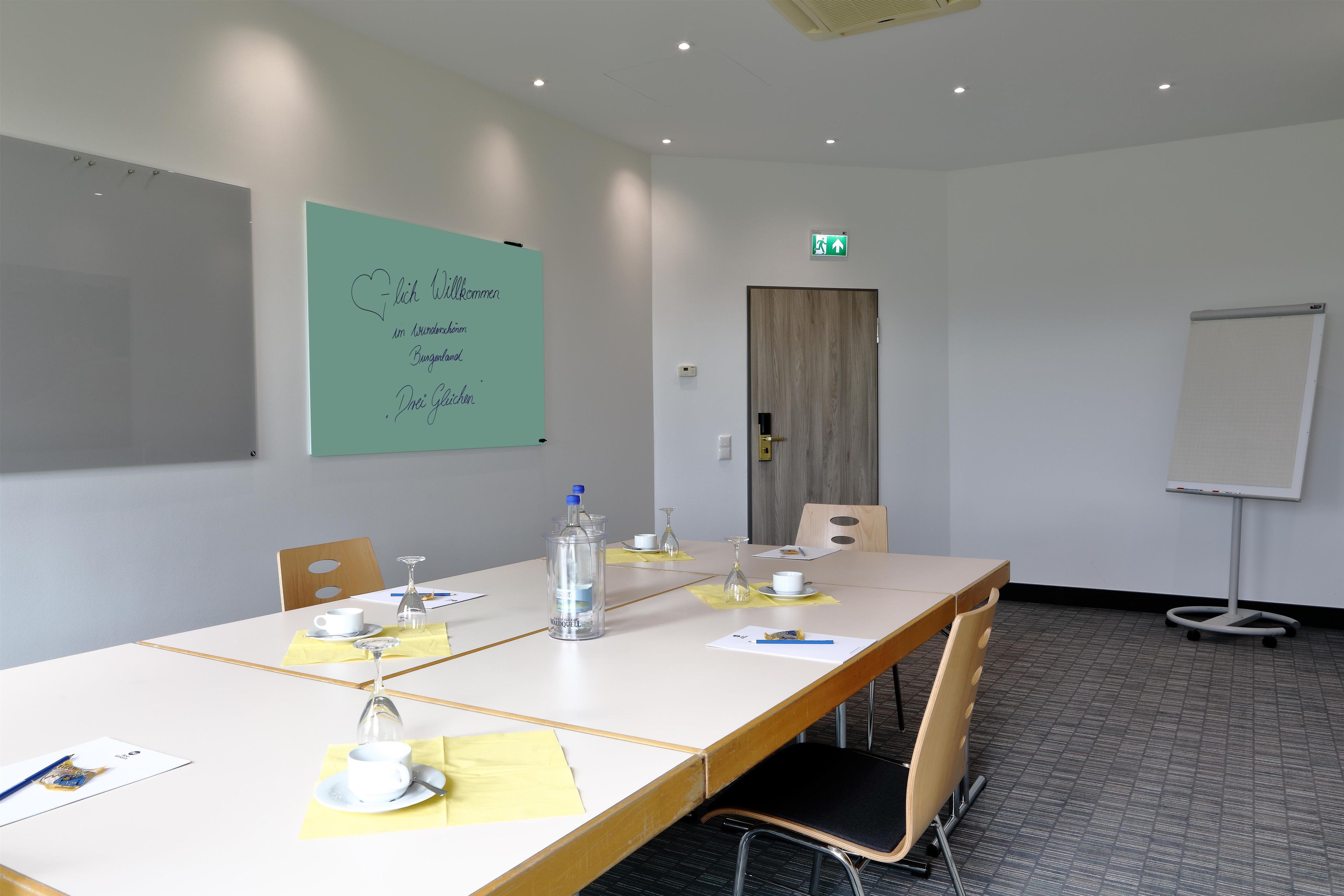 Une salle de conférence avec une longue table, des chaises, un tableau blanc et une porte portant un panneau indiquant la sortie de secours.