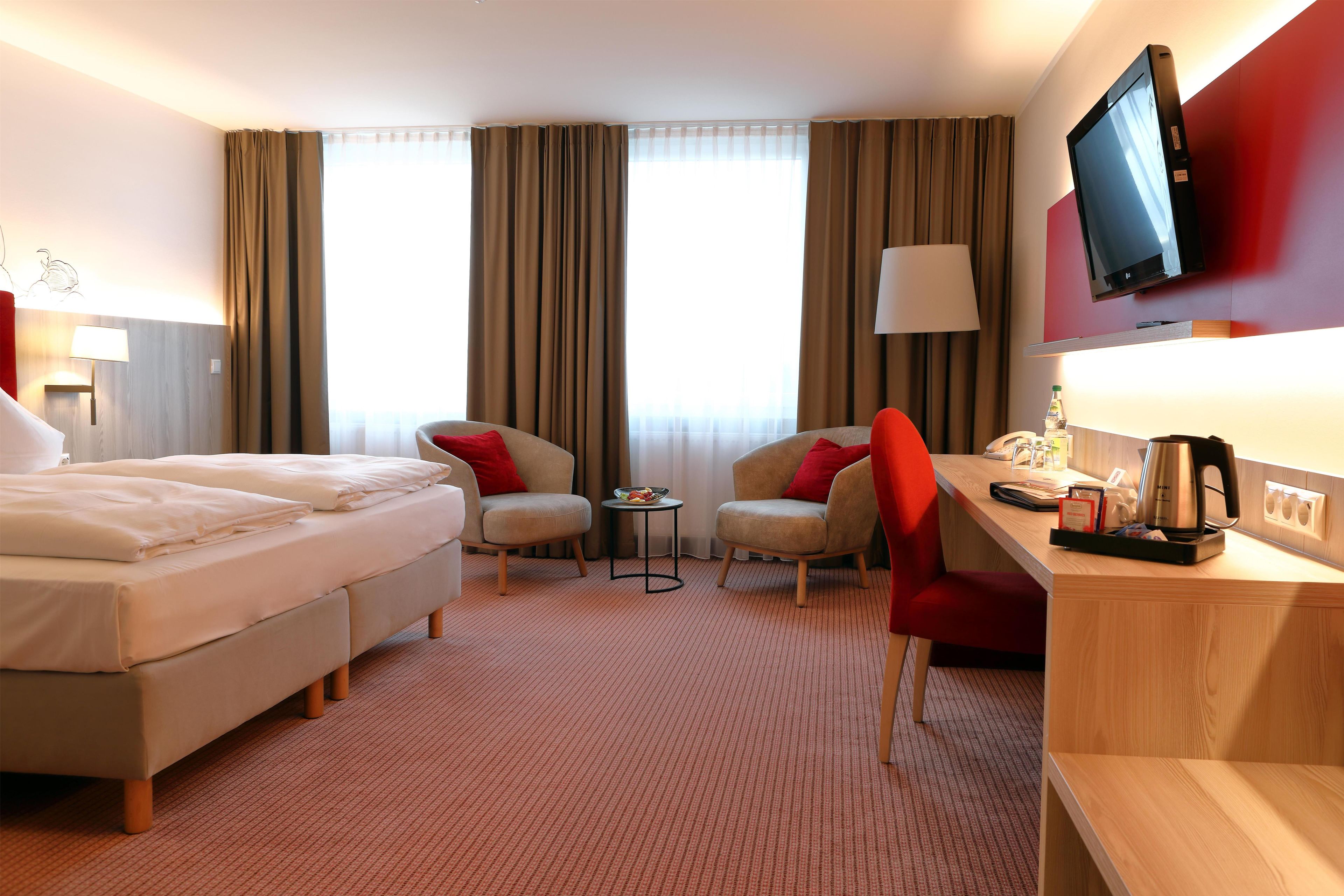 A hotel room with a bed, two armchairs, a desk, a television, and a window with curtains.