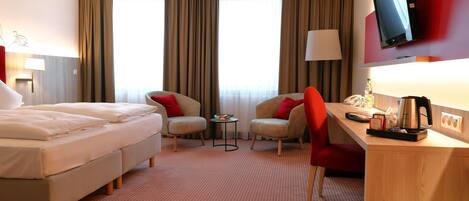 Ein Hotelzimmer mit einem Bett, zwei Sesseln, einem Schreibtisch, einem Fernseher und einem Fenster mit Vorhängen.