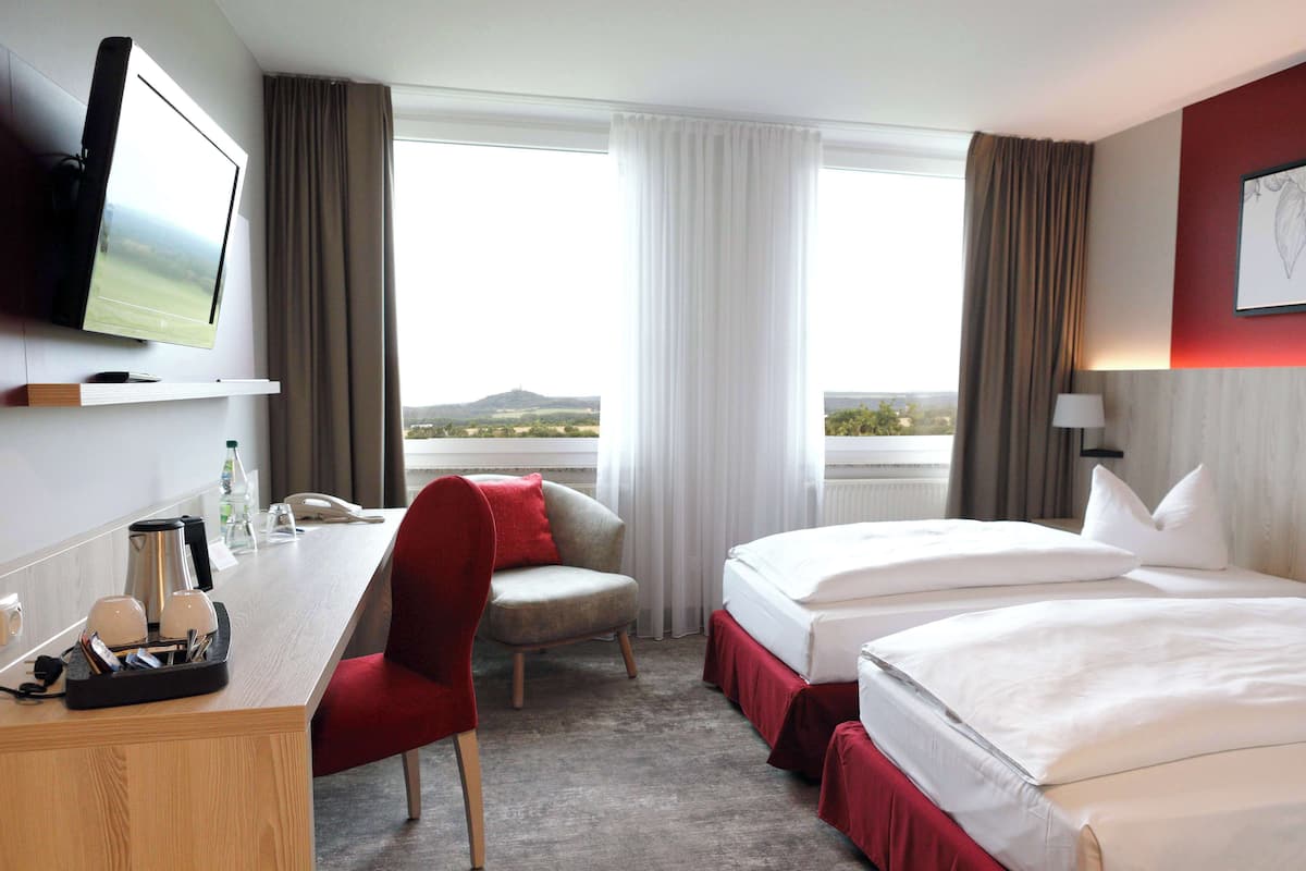 A hotel room with a desk, two beds, a chair, a TV, and a window with curtains.