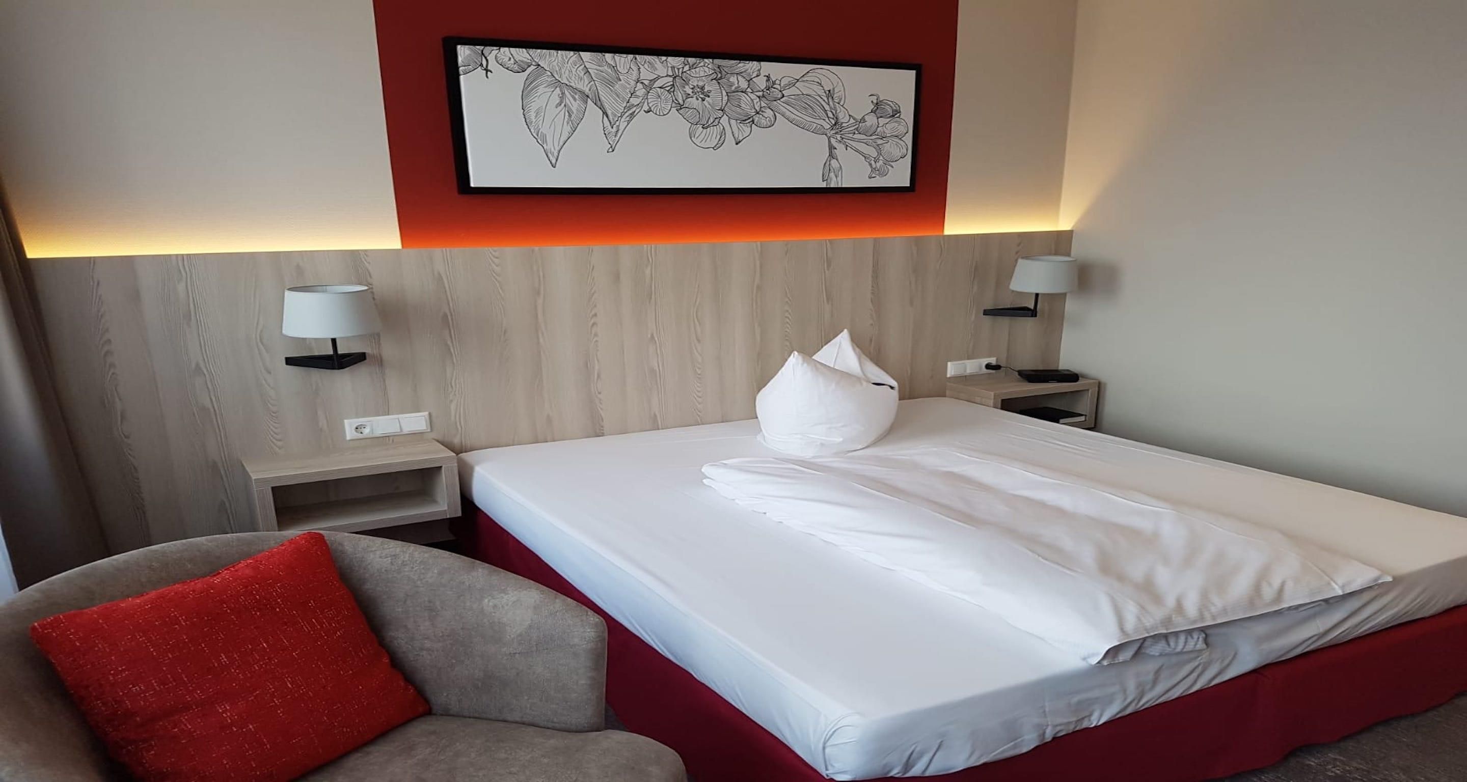 A hotel room with a bed, a red accent wall, a framed botanical illustration, bedside lamps, and a red cushion on a chair.