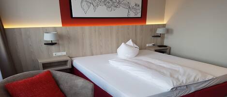 A hotel room with a bed, a red accent wall, a framed botanical illustration, bedside lamps, and a red cushion on a chair.