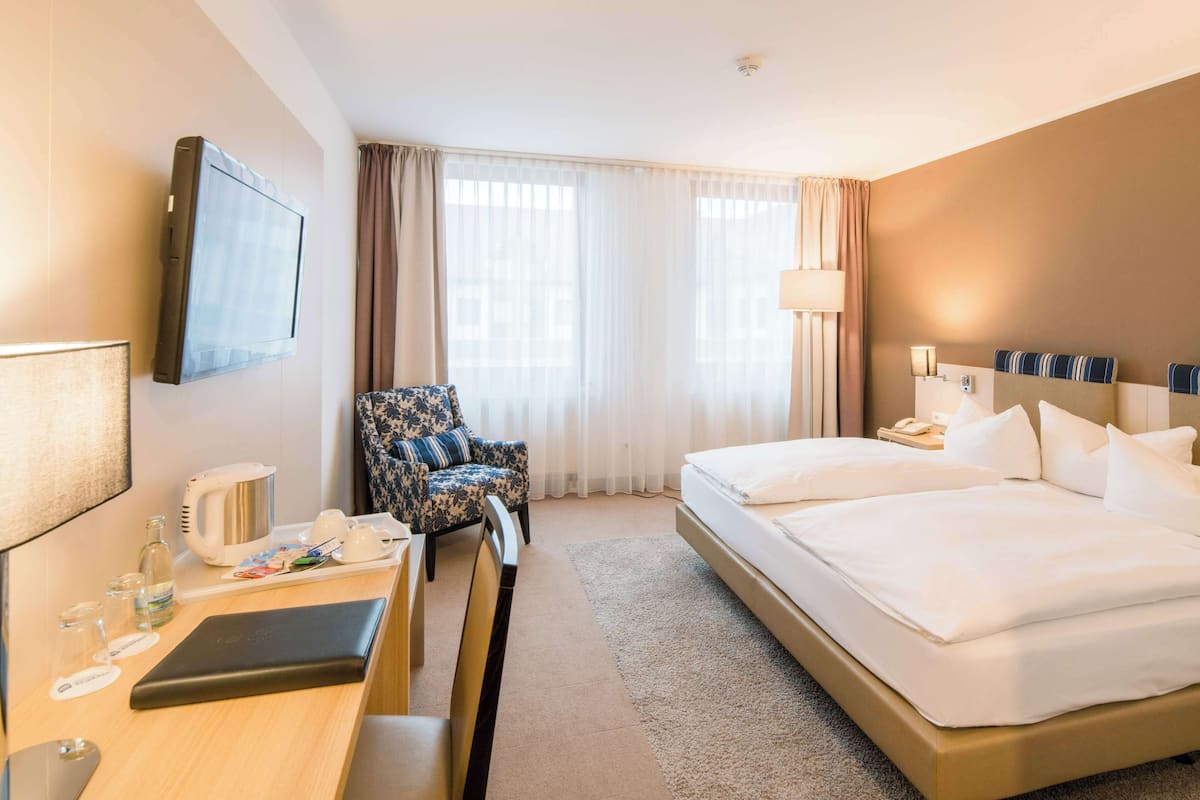A hotel room with a bed, a desk with a laptop, a chair, a TV, and a window with curtains.