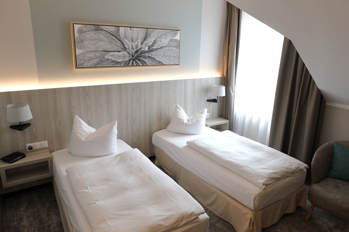 Habitación de hotel con dos camas, una mesita de noche con lámpara, una obra de arte en la pared y vistas al exterior.
