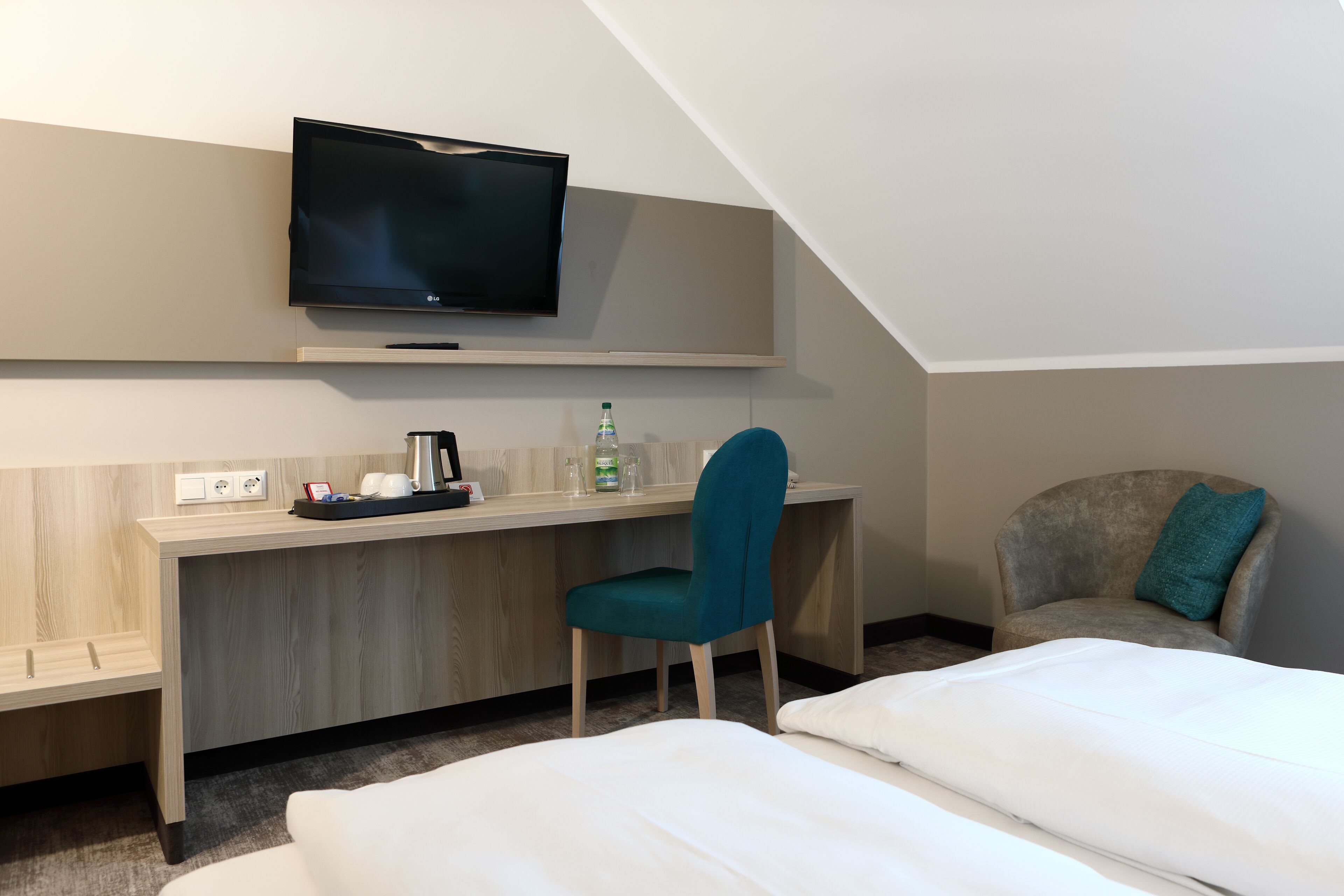 A hotel room with a bed, a desk with a chair, a television, and a small table with a bottle and glasses.