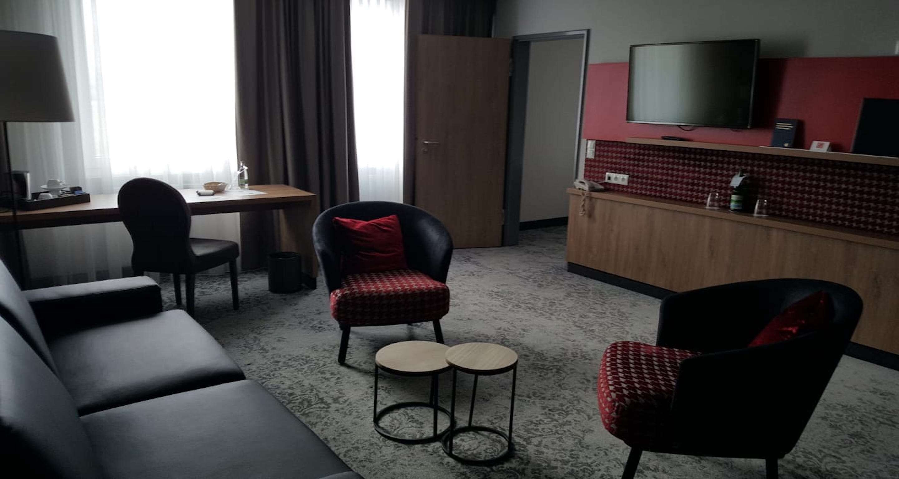 Habitación de hotel con sofá, dos sillones, una mesita, televisor y un área de kitchenette.