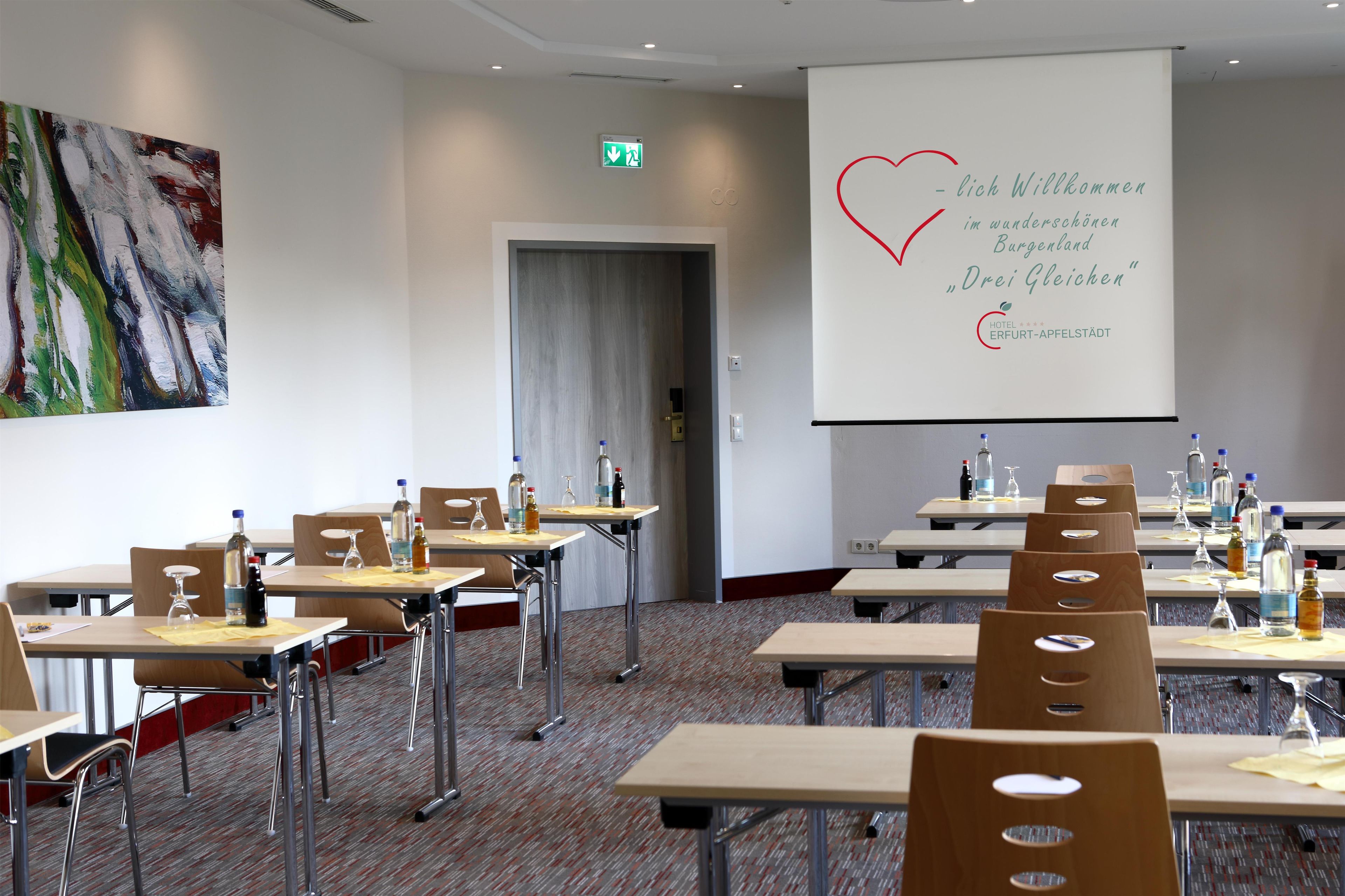 Ein Konferenzraum mit Tischen, Stühlen und einer Projektionsleinwand, auf der eine Begrüßungsnachricht angezeigt wird.