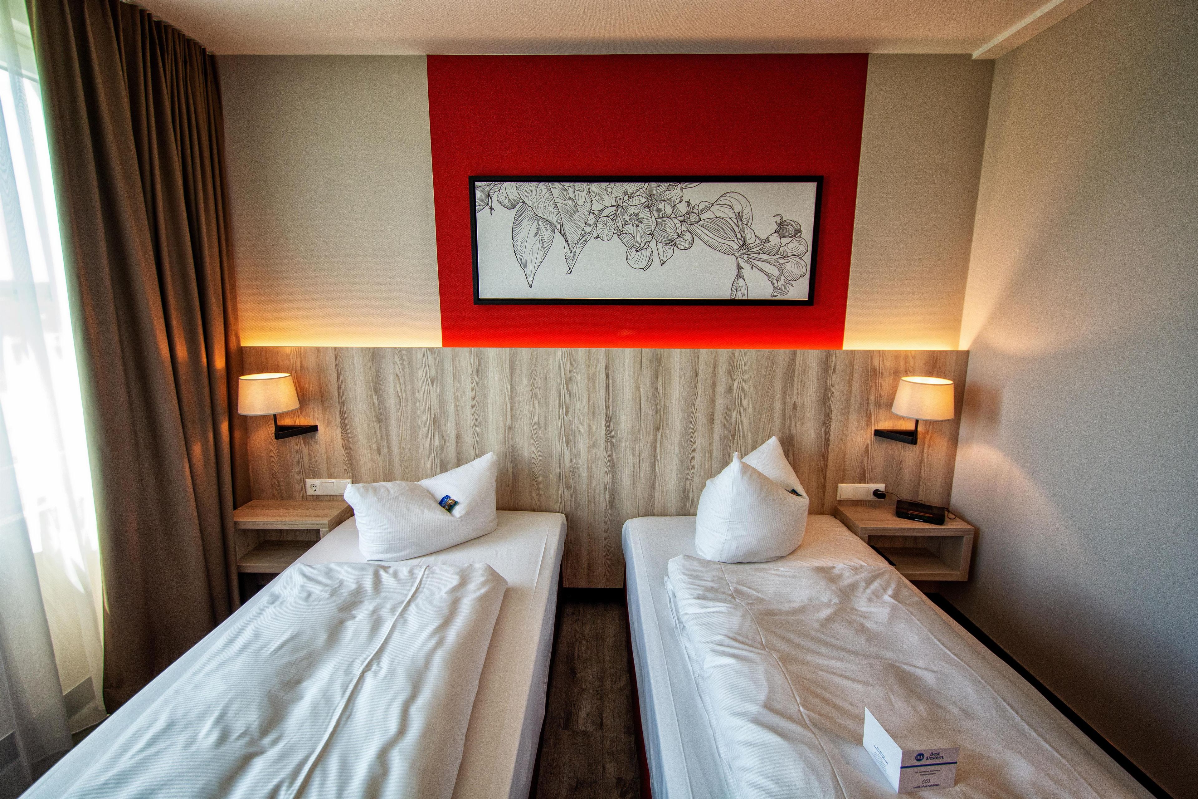 A hotel room with two single beds, a red accent wall, a framed botanical illustration, and bedside lamps.
