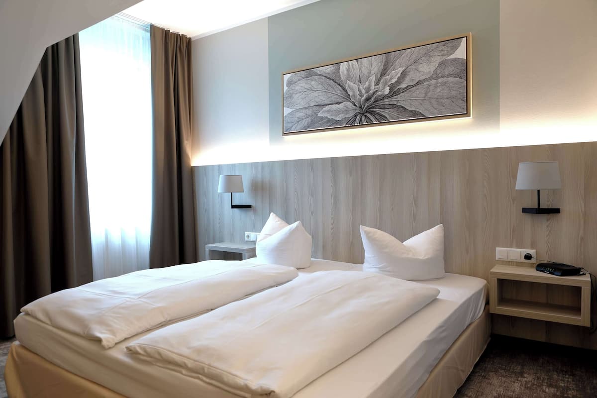 A hotel room with a large bed, two bedside tables, a wall-mounted lamp, and a framed artwork above the bed.
