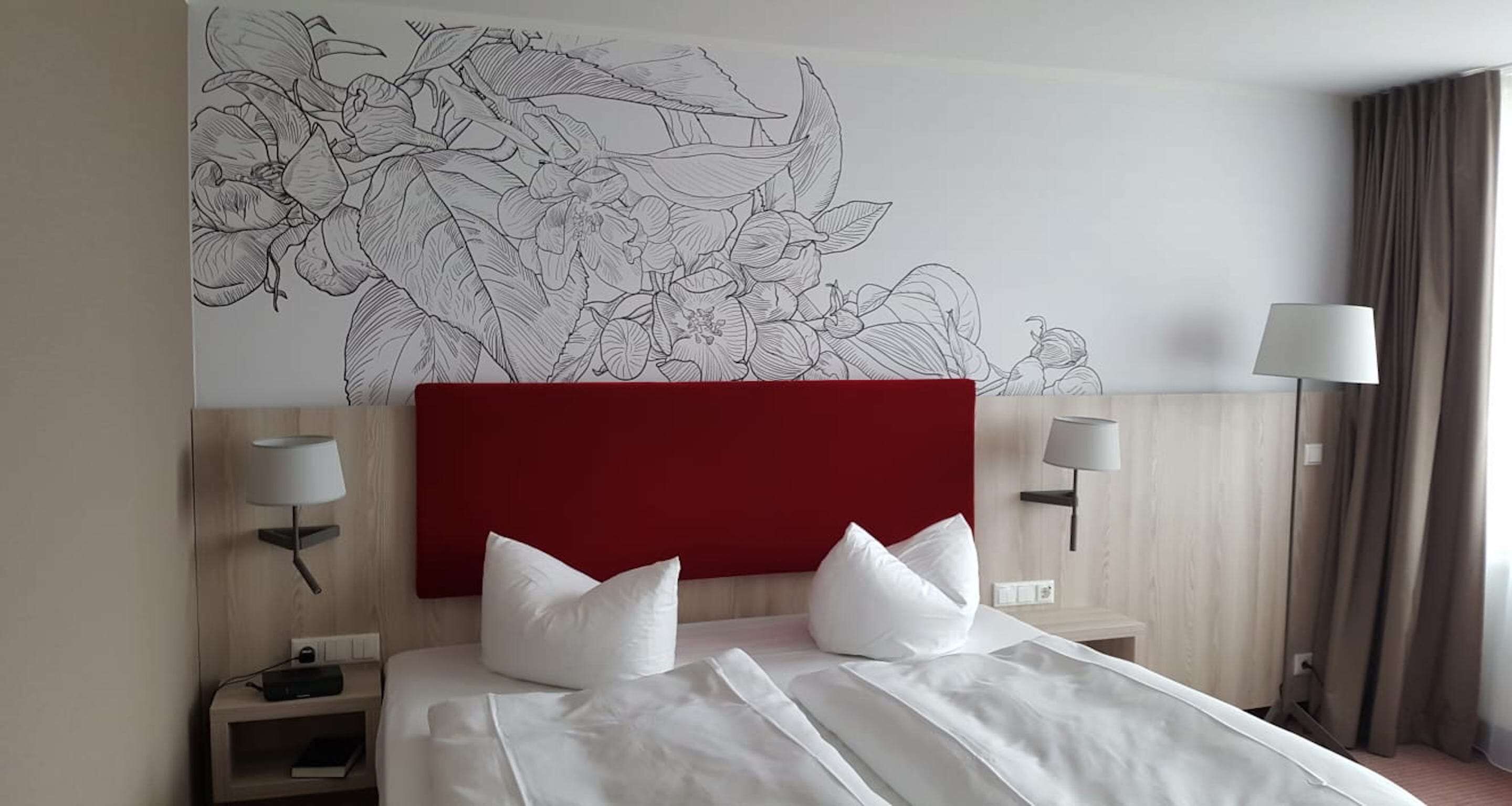 A hotel room with a large floral mural on the wall, a red headboard, two bedside lamps, and a neatly made bed with white linens.